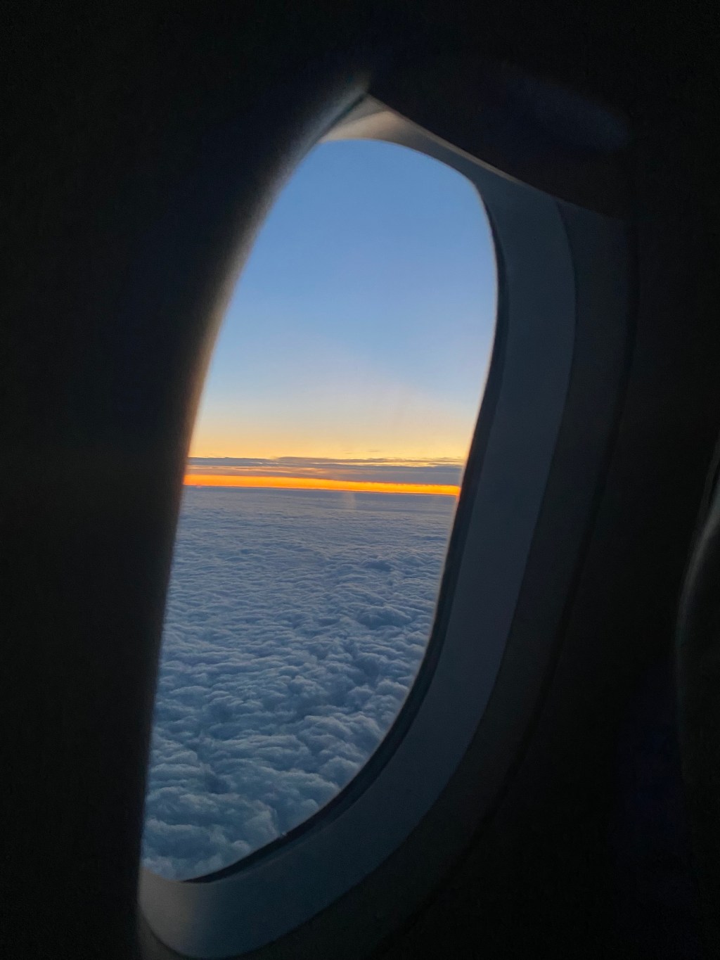 View from an airplane window above a thick blanket of clouds at sunrise, with a thin band of glowing orange light along the horizon and a soft blue sky fading into darkness inside the cabin.