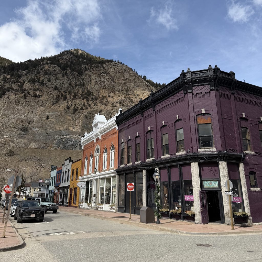 Colorful historic storefronts line a quiet small-town street at the base of a rocky mountain, with a prominent dark purple corner building in the foreground, parked cars along the road, and a bright blue sky with scattered clouds overhead.