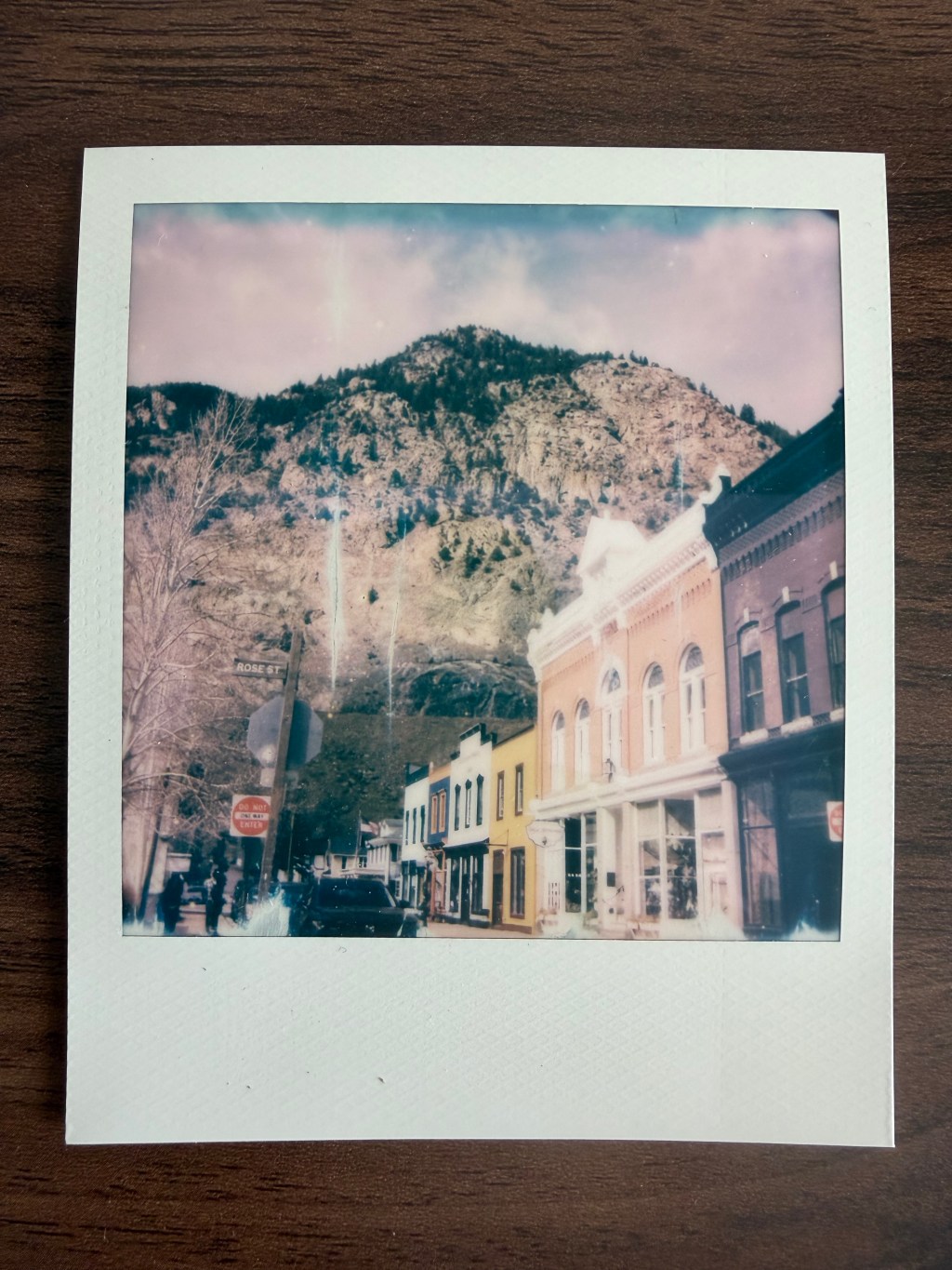 A Polaroid-style photograph resting on a wooden table shows a small-town street with colorful historic storefronts and parked cars, set beneath a rocky mountain covered with sparse trees. A street sign reading “Rose St” is visible near a stop sign, and the photo has a slightly faded, vintage look with light streaks. 📷⛰️