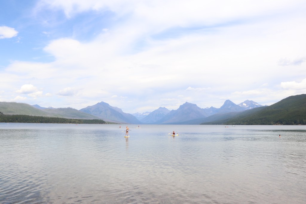 wide scenic mountain range in the background as people on paddleboards float on a crystal-clear lake nearby.