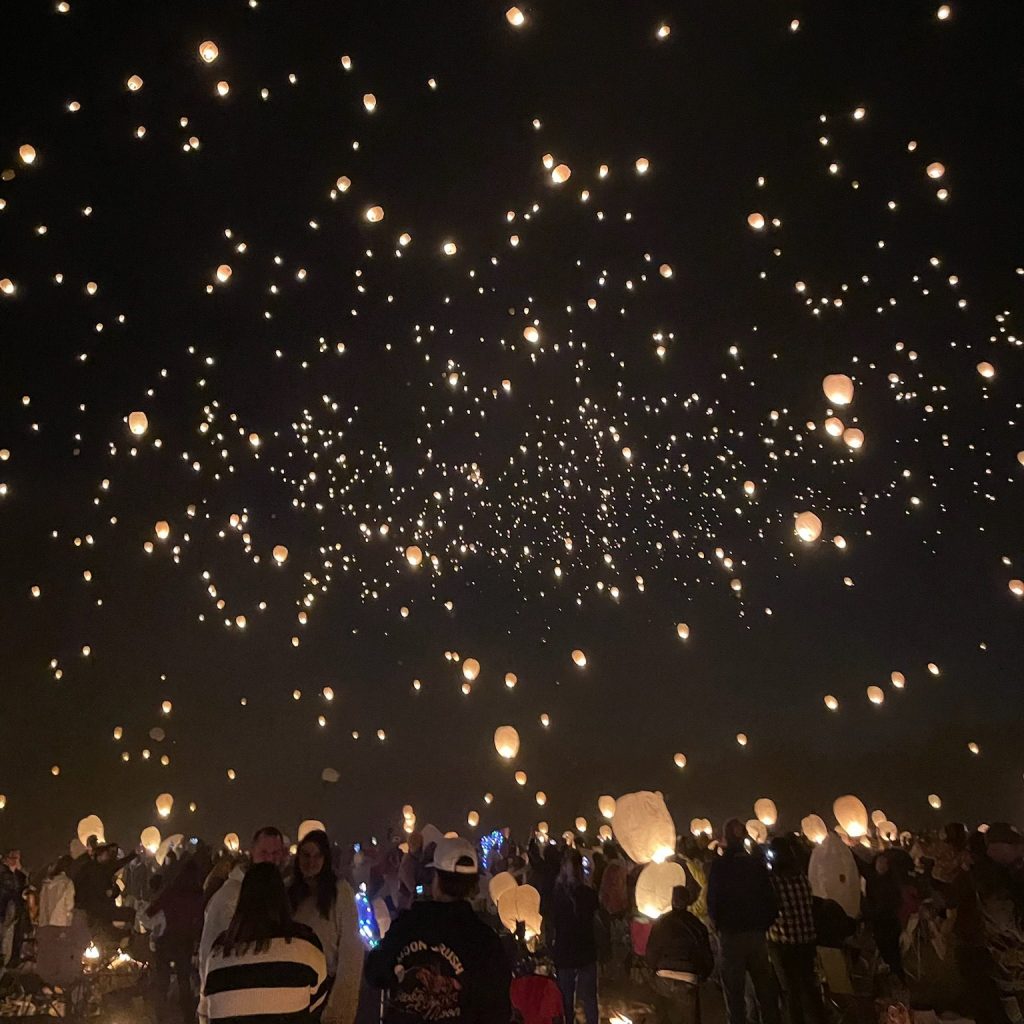 Large crowd gathered at night watching hundreds of glowing sky lanterns rise into the dark sky, creating a stunning display of warm lights floating upward like stars.