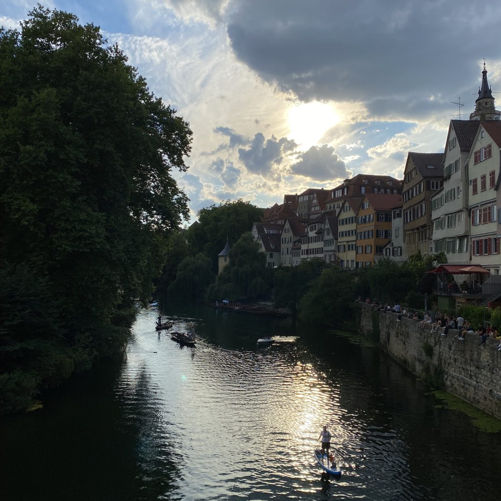Scenic view of a European riverside town at sunset, with colorful historic buildings lining the water, people gathered along a stone embankment, and several boats and a paddleboarder gliding across the river beneath a dramatic sky with sunlight breaking through clouds.