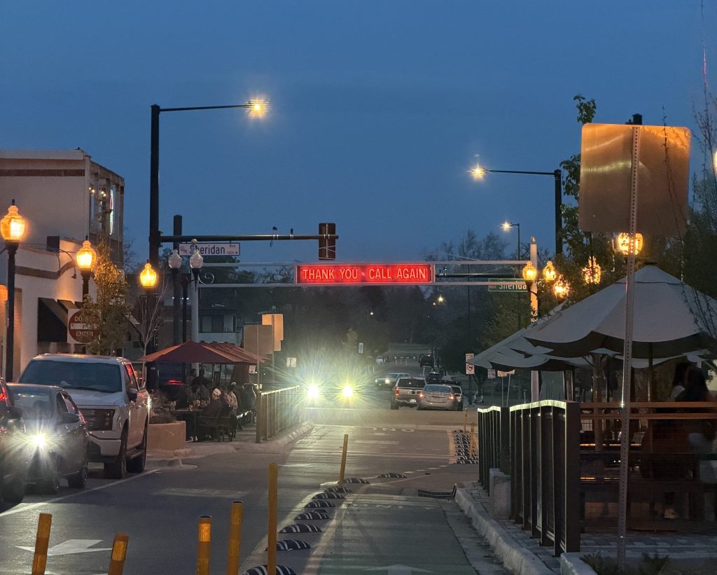 Evening street scene in a small town with glowing streetlights, cars driving past outdoor dining areas, and a lit sign overhead reading “THANK YOU, CALL AGAIN,” under a dusky blue sky with faint lights in the distance.