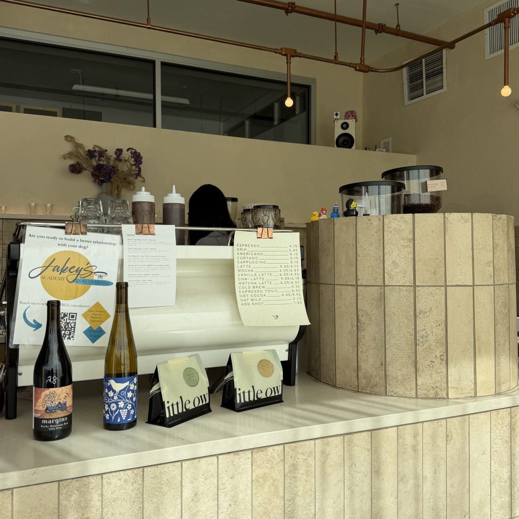 Minimalist coffee shop interior with cream-tiled counters, exposed copper pipe lighting, and hanging bare bulbs. On the counter are wine bottles, coffee bags, and a small espresso menu, with large coffee bean containers to the right. A barista stands behind the espresso machine beneath a high ceiling and modern industrial decor.