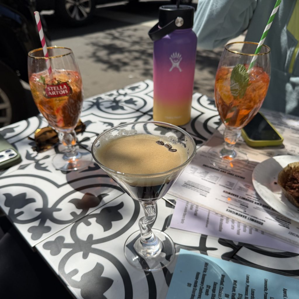Outdoor café table with cocktails and an espresso martini in bright afternoon sunlight, featuring colorful drinks, striped paper straws, menus scattered across the table, and a gradient Hydro Flask water bottle in the center.