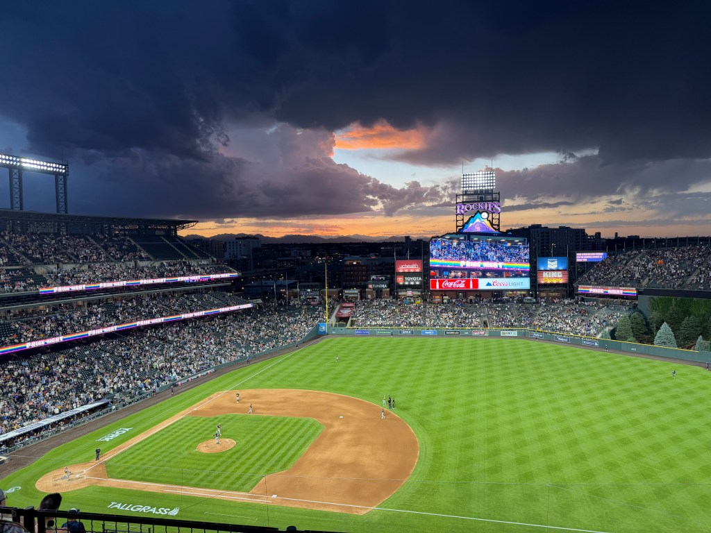 A wide view of a baseball stadium during a game at dusk, seen from high in the stands behind home plate. The field is brightly lit and surrounded by a large crowd. Dark, dramatic storm clouds hang over the stadium, while a break in the clouds reveals a glowing orange and pink sunset above the distant mountains. A large scoreboard in center field displays colorful graphics, creating a striking contrast between the illuminated ballpark and the stormy sky.