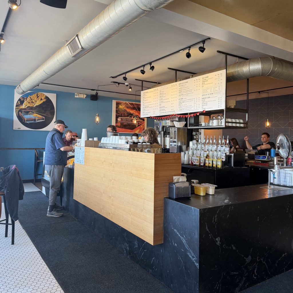 Modern coffee shop interior with a minimalist wood-and-black counter, exposed metal air ducts, and large menu boards overhead. Customers wait and chat while baristas prepare drinks behind the counter, with colorful wall art and warm pendant lighting creating a bright and welcoming atmosphere.
