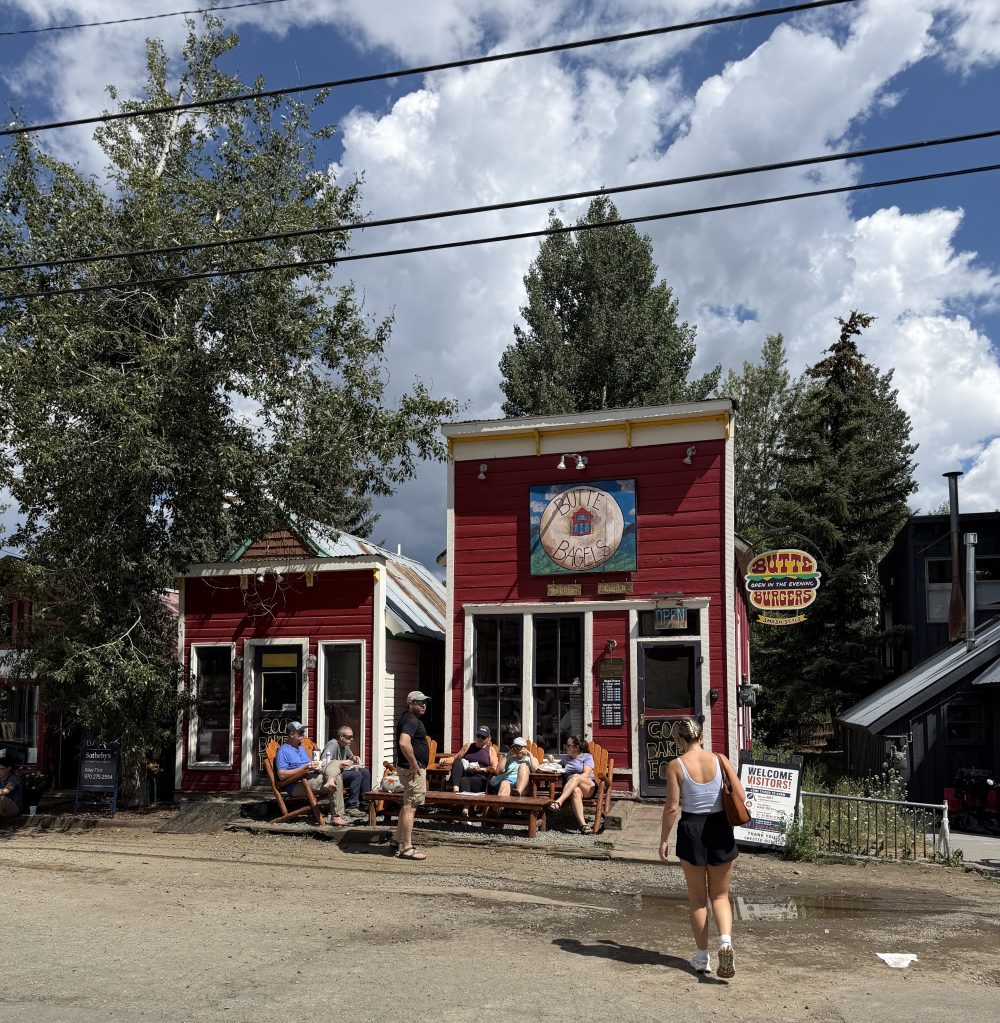woman walks towards a two-story red building reading 'butte bagels' in Crested Butte colorado under a cloudy blue sky.
