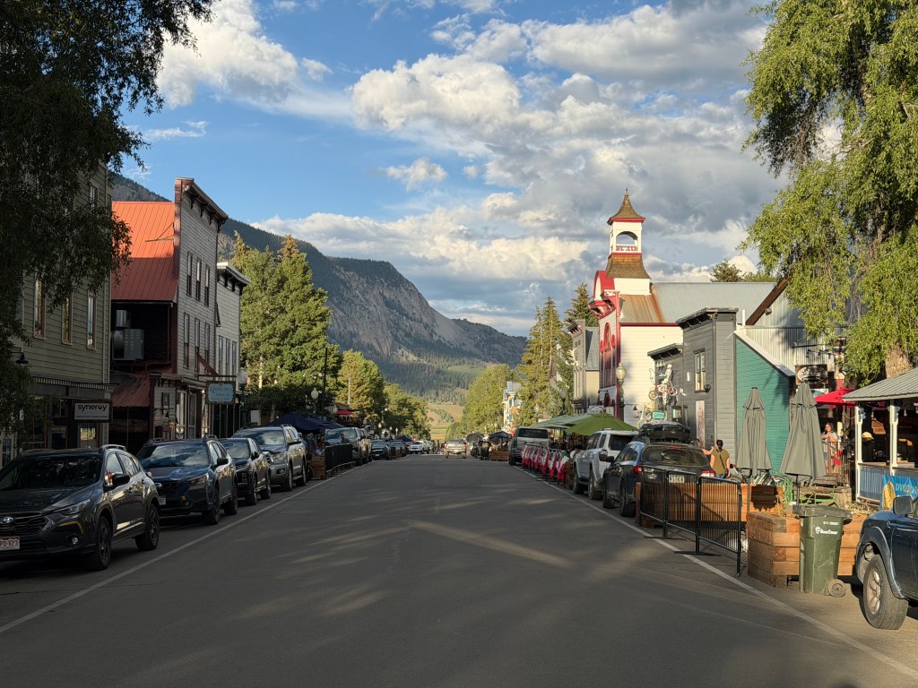 the quiet main street of crested butte, colorado rests at the base of crested butte mountain under a partly cloudy blue sky.